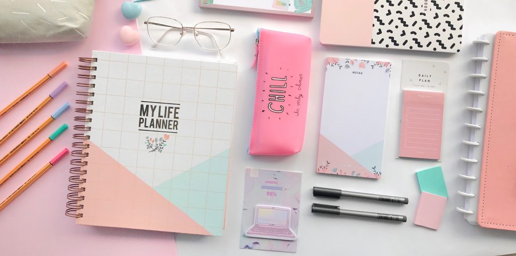 pink notebook beside black click pen and black framed eyeglasses